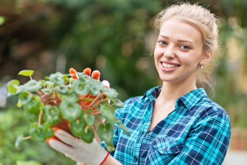A young woman with blonde hair tied back, wearing a blue and green checked shirt and gardening gloves, is standing outdoors in a garden setting. She is holding a small potted plant with green, lobed leaves and some pinkish flower buds. The background features a neatly maintained garden with various plants and blurred foliage, suggesting a lush, well-kept outdoor space typical of a residential garden in Kensington. The scene is lit with natural daylight, indicating a clear, sunny day. The woman appears to be engaging in gardening activities such as planting or tending to her garden, reflecting professional garden maintenance or personal gardening efforts. This image aligns with landscaping and gardening services provided by Gardening Services Kensington, emphasizing attention to detail and care in outdoor garden spaces in the Kensington area.