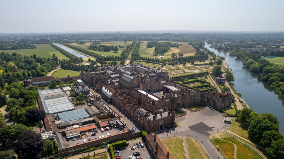 An aerial view of the gardens surrounding a historic castle-like structure, with well-maintained lawns, neatly trimmed hedges, and garden beds. The grounds feature pathways and gravel areas, alongside
