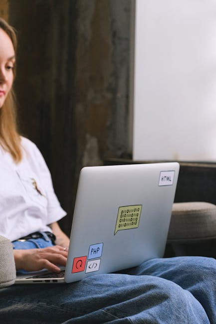 A person wearing a white shirt and blue jeans is sitting outdoors next to a garden, using a silver laptop placed on their lap. The laptop has various colorful stickers, including a yellow one with bin